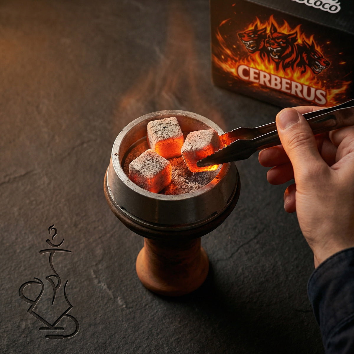 Glowing Cerberus Coco 26mm coconut shisha charcoal cubes in a heat management device on a hookah, hand with tongs placing a coal with the lounge box in the background., image 2 of 11
