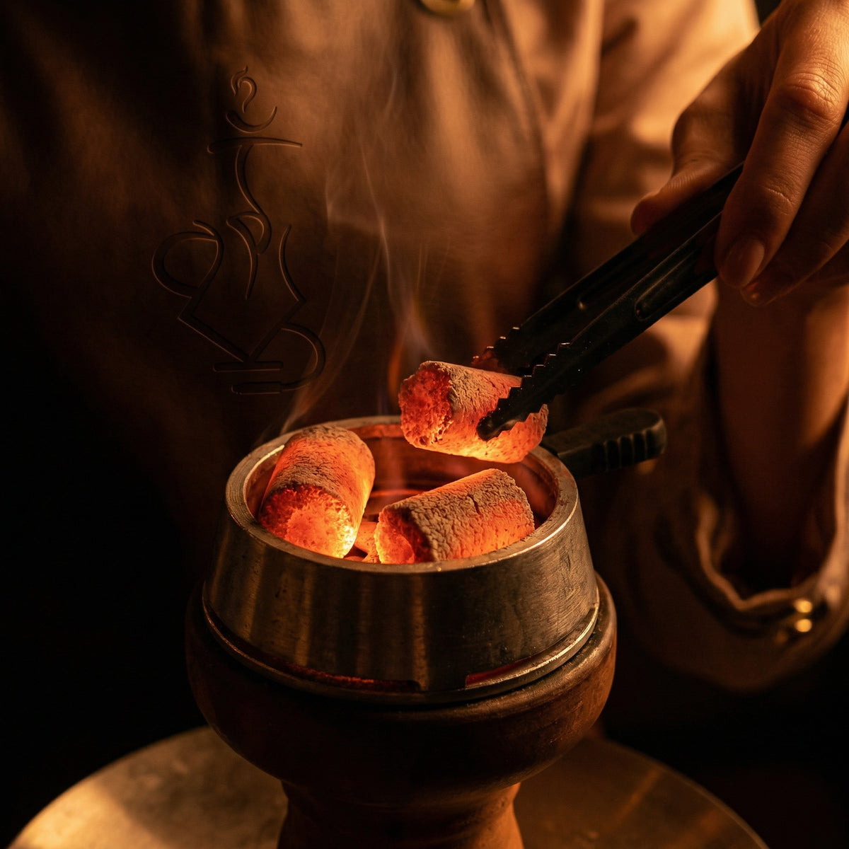 Glowing Cerberus Coco Finger coconut charcoal coals inside a heat management device with a hand placing a coal using tongs during a hookah session, image 2 of 4