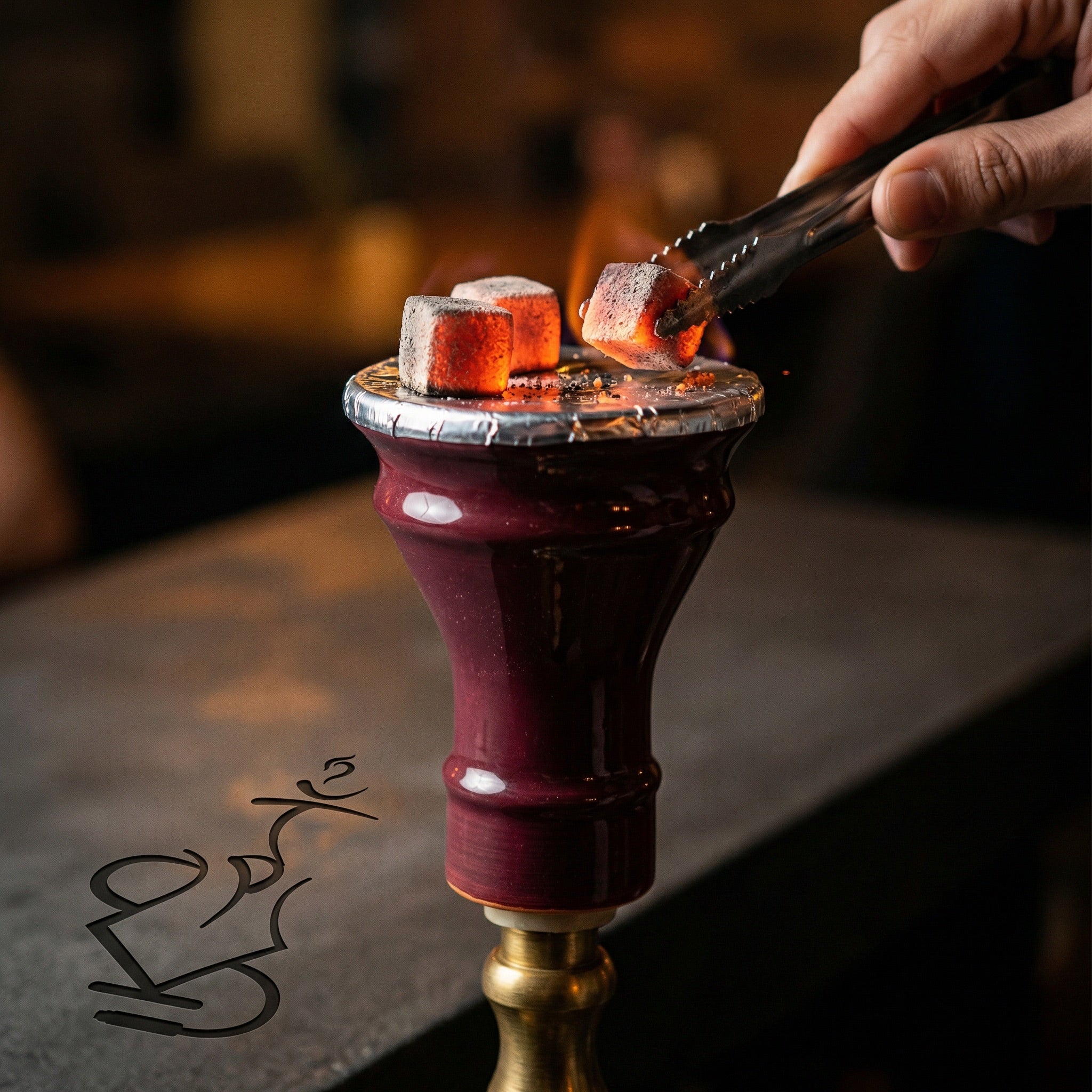 King Buzz Traditional Egyptian Bowl foiled with glowing coals on a hookah, a hand rotating charcoal with tongs in a warm dimly lit session setting.